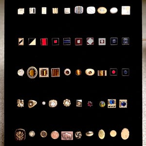 May include: A collection of assorted metal cufflinks arranged in five horizontal rows on a black display board. The cufflinks feature various shapes including squares, rectangles, circles, and ovals, with diverse finishes, enamel inlays, and gemstones.