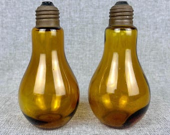 May include: Two amber-colored glass bottles shaped like light bulbs. Each bottle has a brown metal base. The bottles are displayed on a gray surface. The glass has a glossy finish.