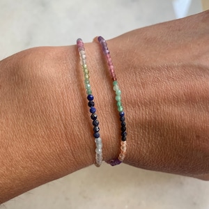 May include: Two beaded bracelets with colorful gemstones. The bracelets are layered on a person's wrist. The top bracelet features pink, green, and blue gemstones. The bottom bracelet features light blue, green, and dark blue gemstones.