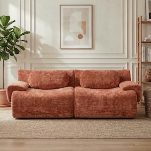 May include: A two-piece, coral-colored sofa with rounded arms and back cushions. The sofa sits on a neutral-toned rug, with a potted plant and framed artwork in the background. The sofa's plush texture and color create a cozy aesthetic.