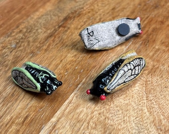 Puede incluir: Tres figuras de cerámica de cigarras. Una es de color gris claro con un imán negro, otra es negra con alas color canela y ojos rojos, y la tercera es verde y negra. Las figuras están sobre una superficie de madera.