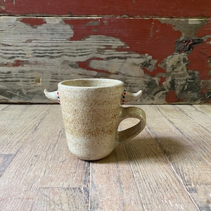 May include: A speckled white ceramic mug with red and blue accents on the handles. The mug has a rounded shape and a handle on the side.