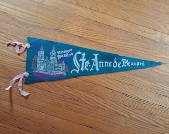 Könnte beinhalten: Ein grünes Wimpel mit dem Text "Ste. Anne de Beaupré" in weißer Schrift. Das Wimpel zeigt eine Illustration einer Kirche mit zwei Türmen und den Worten "Basilique Basilica". Rosa Bänder sind am Wimpel befestigt.