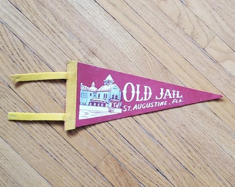 May include: A maroon pennant with the words "OLD JAIL ST. AUGUSTINE, FLA." and an illustration of a building. The pennant has yellow trim and two yellow tabs for hanging.