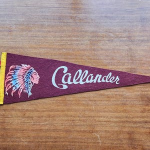 Op de afbeelding: Bordeaux vilten wimpel met het woord "Callander" in wit script. Een Indiaans profiel met een gevederde hoofdtooi staat links. De wimpel heeft een gele rand en rode lintlipjes. De wimpel ligt op een houten oppervlak.