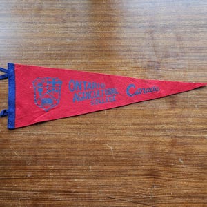 May include: A red felt pennant with a blue vertical strip on the left side featuring two small ties. The pennant displays a blue crest, the text "ONTARIO AGRICULTURAL COLLEGE", and the word "Canada" printed in blue ink across the centre surface.