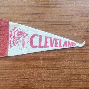May include: A triangular felt pennant in off-white with a red vertical strip on the left side. The pennant features a red graphic of a person wearing a feathered headdress and the red text CLEVELAND printed in a bold, sans-serif capital font.