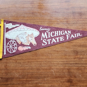 May include: A maroon pennant with the text "Souvenir of Michigan State Fair" in white. The pennant features illustrations of a Ferris wheel, a cow, a horse, and pumpkins. The pennant has a yellow border and a yellow loop for hanging.