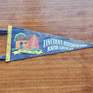 May include: A dark blue felt pennant featuring a graphic of a log cabin and the text "LINCOLN'S BOYHOOD HOME KNOB CREEK, KY." and "1811 TO 1816" on a yellow banner. The pennant has a yellow vertical strip on the left side with two blue ties.