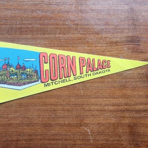 Könnte beinhalten: Ein gelbes Wimpel mit den Worten "CORN PALACE MITCHELL, SOUTH DAKOTA" in orangefarbenen Buchstaben. Der Wimpel zeigt eine Illustration eines Gebäudes mit roten Kuppeln und Flaggen vor blauem Himmel.
