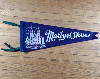 Könnte beinhalten: Ein blaues Filz-Wimpel mit weißem Text, der "Martyrs' Shrine Midland Canada" lautet. Das Wimpel zeigt eine weiße Illustration eines Kirchengebäudes.