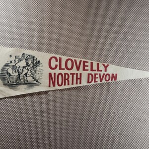 Peut inclure: Un fanion triangulaire en feutre blanc cassé présentant une illustration à l'encre noire de deux ânes près d'un bâtiment en pierre. Le texte CLOVELLY NORTH DEVON est imprimé en lettres capitales rouges sur le tissu.