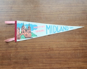 May include: A triangular felt pennant featuring a light blue background with a graphic illustration of the Martyrs Shrine in Midland, Ontario. The text reads Martyrs Shrine Midland Ont in blue and pink lettering, with two pink fabric ties on the left.