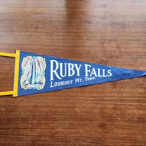 Peut inclure: Un fanion en feutre bleu orné d'une illustration graphique d'une cascade dans un cadre décoratif, avec le texte blanc "RUBY FALLS" et "LOOKOUT MT., TENN." imprimé. Le fanion possède une bande verticale jaune avec deux attaches jaunes.