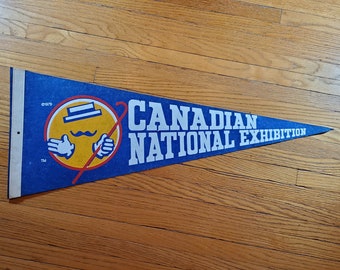 May include: A blue pennant featuring a yellow cartoon character wearing a hat and holding a cane. The text "CANADIAN NATIONAL EXHIBITION" is printed in white on the pennant. The copyright date "C1979" is printed in the top left corner.