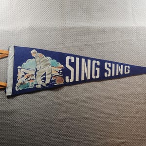 May include: A vintage blue pennant with the text "SING SING" in white block letters. The pennant features an illustration of a person in a striped uniform and a brown ball. The words "ALMA MATER" are also visible.