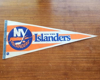 May include: A triangular felt pennant featuring the New York Islanders hockey team logo in blue and orange. The design includes the team name in blue text, a circular logo with a hockey stick and puck, and a small NHL shield logo at the base.