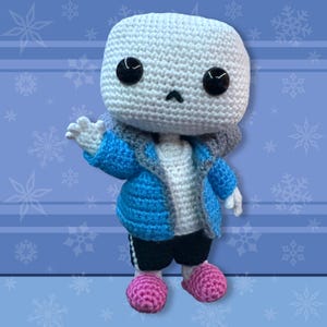 May include: A crocheted doll of a character with a white head, black eyes, and a small black mouth. The doll wears a blue jacket, white shirt, black shorts, and pink shoes. The background is blue with snowflake designs.