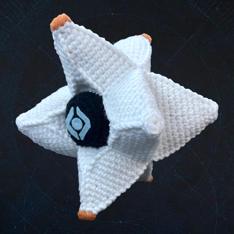 Pode incluir: Um brinquedo de pel&uacute;cia branco em forma de estrela feito de croch&ecirc; com um design de olho preto e azul. O brinquedo tem detalhes laranja nas pontas da estrela.