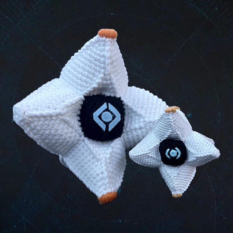 Pode incluir: Duas figuras de Ghost em forma de estrela branca de croch&ecirc; do videogame Destiny. O Ghost maior tem um olho preto e azul, enquanto o Ghost menor tem um olho preto e azul claro.