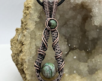 May include: A copper wire wrapped pendant with a green and white agate stone. The pendant is hanging from a black cord and is displayed on a rock.