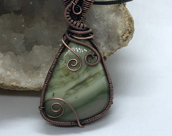 May include: A copper wire wrapped pendant featuring a green and white agate stone. The pendant is attached to a black cord.