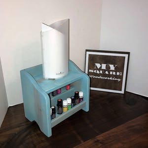 Può includere: Un ripiano in legno blu chiaro con due ripiani per riporre bottiglie di olio essenziale. Il ripiano è posto su una superficie in legno scuro. Un diffusore di olio essenziale bianco è posto sul ripiano superiore. Un cartello in legno con il testo "MY SQUARE Woodworking" è sullo sfondo.