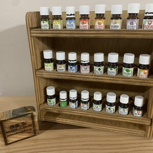 May include: Wooden shelf unit with three tiers, each holding rows of small glass bottles with labels. The bottles are filled with essential oils.