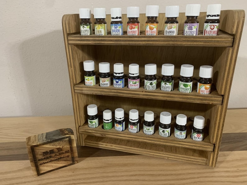 May include: Wooden shelf unit with three tiers, each holding rows of small glass bottles with labels. The bottles are filled with essential oils.