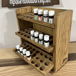 May include: Wooden essential oil rack with three shelves and a pull-out drawer. The rack is made of light brown wood and has a natural wood finish. The rack is filled with small glass bottles of essential oils.