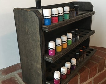 May include: A dark stained wooden essential oil rack with three shelves. The rack is filled with small glass bottles of essential oils in various colors. The bottles are arranged in rows on the shelves.
