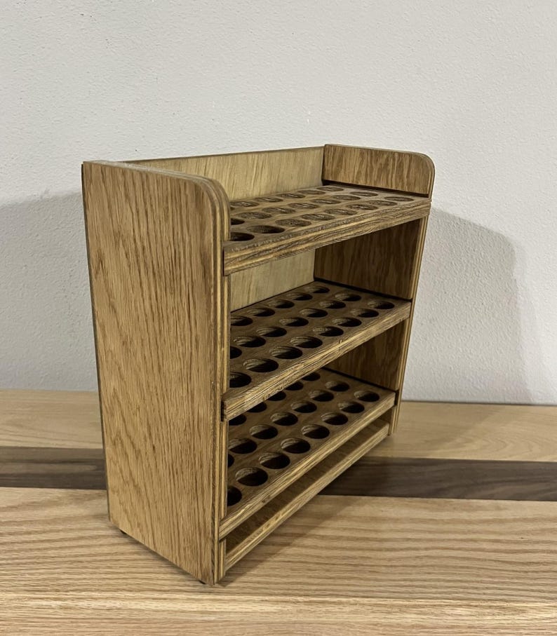 May include: A wooden shelf unit with three tiers, each tier has multiple circular cutouts for holding small bottles or tubes. The shelf is made of light brown wood with a natural wood grain.