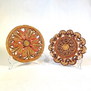 Puede incluir: Dos posavasos de paja trenzada con diseños intrincados. Un posavasos presenta un gran diseño floral en tonos marrones, naranjas y rosas. El otro posavasos tiene un diseño circular con remolinos en tonos marrones y rosas.
