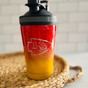 May include: A red and yellow stainless steel shaker bottle with a black lid and a silver band. The bottle has a Kansas City Chiefs logo on the side. "SHAK" is printed on the lid.