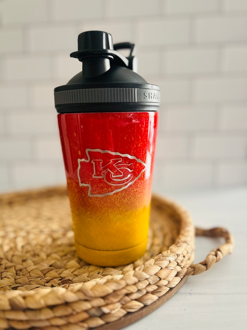 May include: A red and yellow stainless steel shaker bottle with a black lid and a silver band. The bottle has a Kansas City Chiefs logo on the side. "SHAK" is printed on the lid.
