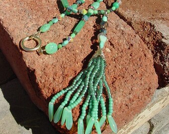 Op de afbeelding: Een lange groene en gouden ketting met een kwast. De ketting is gemaakt van kralen en heeft een gouden sluiting. De kwast is gemaakt van groene kralen en is ongeveer 25 cm lang.