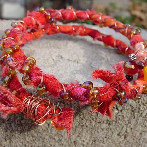 May include: A red fabric bracelet with copper wire and brown glass beads. The bracelet is wrapped around itself and has a yellow star charm.