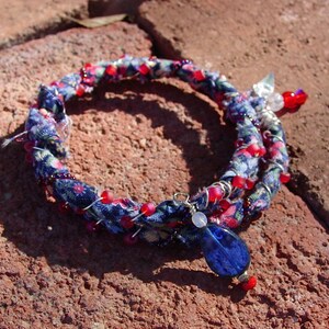 Könnte beinhalten: Ein blaues und rotes Stoffarmband mit einem blauen Glasanhänger. Das Armband besteht aus Stoffresten und Perlen.
