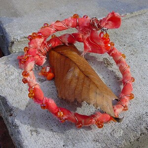 Könnte beinhalten: Ein handgemachtes Armband aus orangefarbenen Perlen und rosa Stoff, der um einen Kupferdraht gewickelt ist. Das Armband liegt auf einer grauen Betonfläche mit einem braunen Blatt.