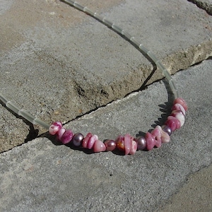 Puede incluir: Un collar con cuentas de piedras preciosas rosas y moradas, intercaladas con pequeñas cuentas redondas de color púrpura oscuro. El collar está ensartado en un cordón transparente segmentado.