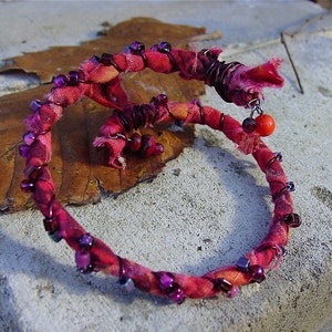 Peut inclure: Un bracelet en tissu rouge et violet avec du fil et des perles. Le bracelet est enroulé autour d'une feuille brune.