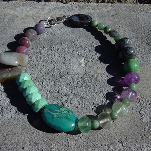 Puede incluir: Un collar de piedras preciosas multicolores con una variedad de piedras, incluyendo verde, morado, marrón y gris. El collar está hecho con un cierre de plata y presenta una piedra turquesa grande de forma ovalada.