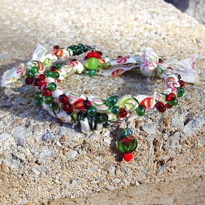 May include: A white fabric bracelet with red, green, and white beads. The bracelet is wrapped around a rough, gray surface.