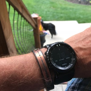 May include: A black wristwatch with a compass, altimeter, and barometer features. The watch face displays the time as 7:18. The watch is worn on a person's wrist with a copper bracelet with a sound wave design.