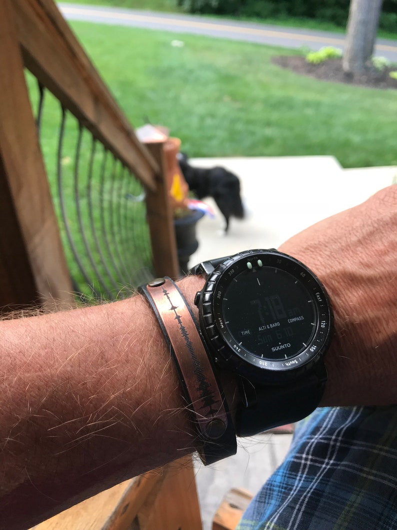 May include: A black wristwatch with a compass, altimeter, and barometer features. The watch face displays the time as 7:18. The watch is worn on a person's wrist with a copper bracelet with a sound wave design.