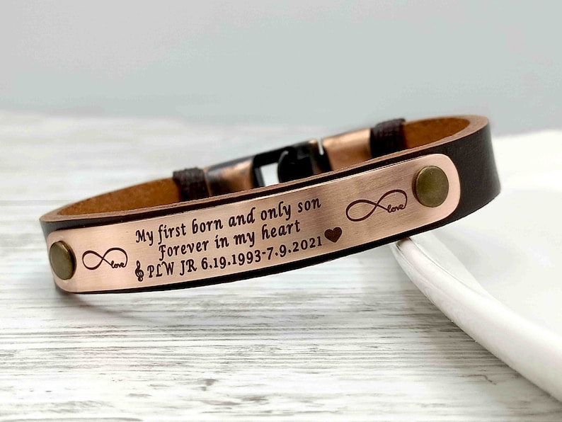 May include: Brown leather bracelet with a rose gold metal plate engraved with the text "My first born and only son Forever in my heart PLW JR 6.19.1993-7.9.2021" and infinity symbols with the word "love" and a heart.