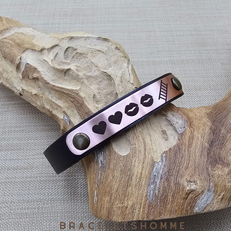 May include: A black leather bracelet with a rose gold metal plate featuring a ladder, hearts, and lips.