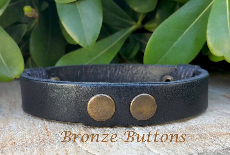 May include: Black leather bracelet with two bronze button accents. "Bronze Buttons" is written in brown text below the bracelet.