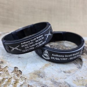 May include: Two black leather bracelets featuring silver-toned metal plates engraved with the names Anthony Scott Walker and CPL Todd Mcgurn, their birth and death dates, crossed rifle icons, and a military-style crest emblem with black studs.