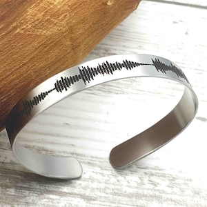Könnte beinhalten: Ein silbernes Armband mit einem schwarzen Schallwellendesign, das auf der Vorderseite graviert ist.
